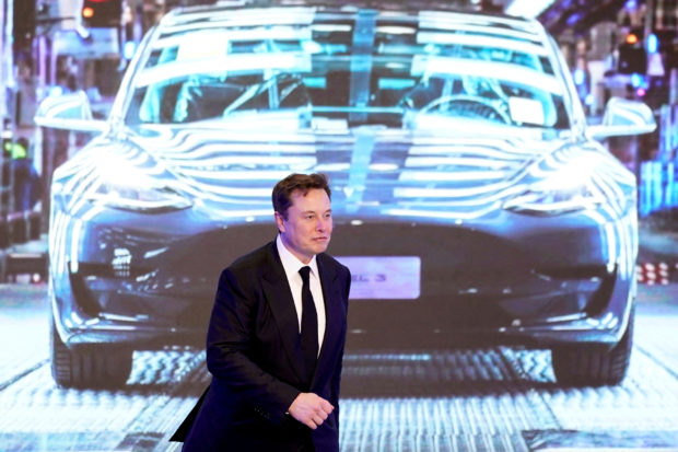 Elon Musk walks next to a screen showing an image of Tesla unit
