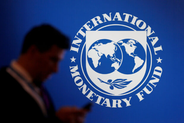 A man checks his phone while standing near a logo of the IMF