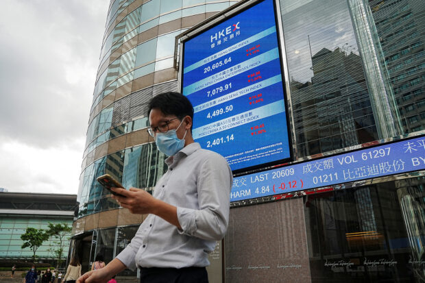 Man checks his phone in front of an electronic screen displaying the Hang Seng stock index