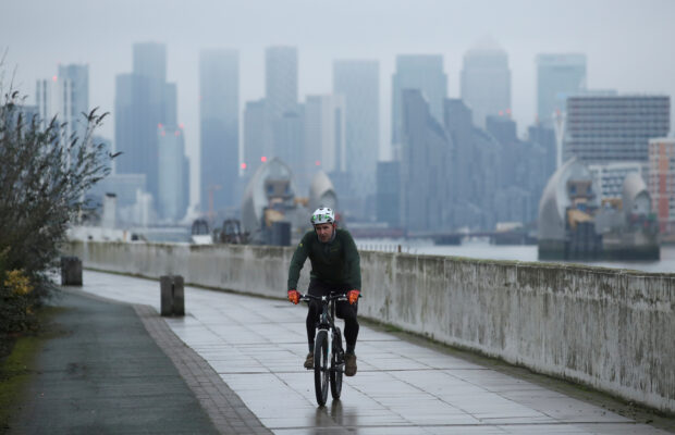 A man cycles along a path in London with the Canary Wharf business district in the background