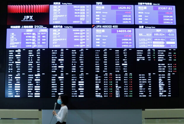 A TV reporter stands in front of a large screen showing stock prices in Tokyo
